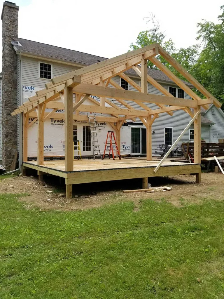 Timber frame addition with gable truss raised on a deck behind a farmhouse